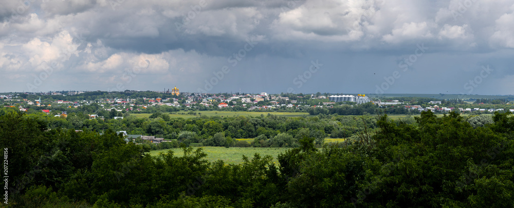 Obraz premium Pidvolochysk, Ternopil Oblast, Ukraine: A panoramic view of the town under dramatic storm clouds, a prominent golden-domed church and modern grain silos amidst vast green landscapes and fields