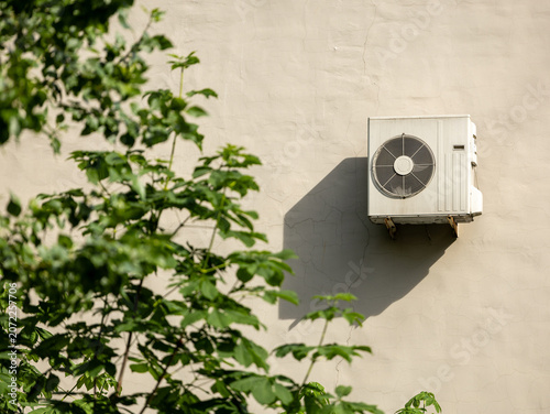 White AC unit mounted on light stucco wall. Green leaves blur foreground. Sharp shadow casts sunlit facade. Contrast between modern utility and fresh spring nature.