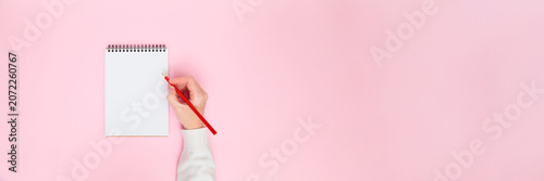 Female hand writing on blank notebook on pastel pink background. Minimal flat lay banner with copy space for notes, planning, education, business, blogging, creative content