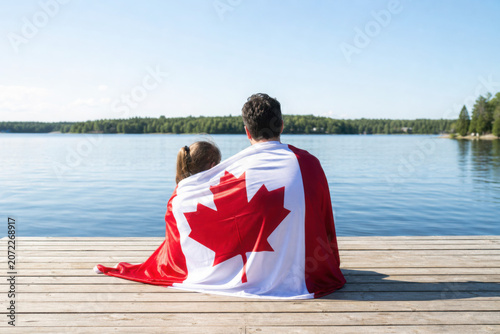 Father and daughter wrapped in large Canadian flag sitting on wooden dock by lake. Canada Day celebration outdoor. Dad and child sitting together on 1 of July celebrating national Canada Day.
