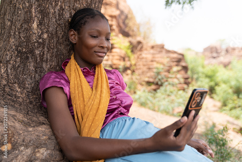 Leaning against the tree, an African girl sits quietly with her phone, lost in a calm moment of reflection beneath the soft light of the afternoon.