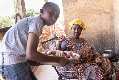 In the warmth of a sunlit courtyard, a mother and son share a quiet bond over daily chores, learning the value of contribution as the family's traditional meal simmers nearby.