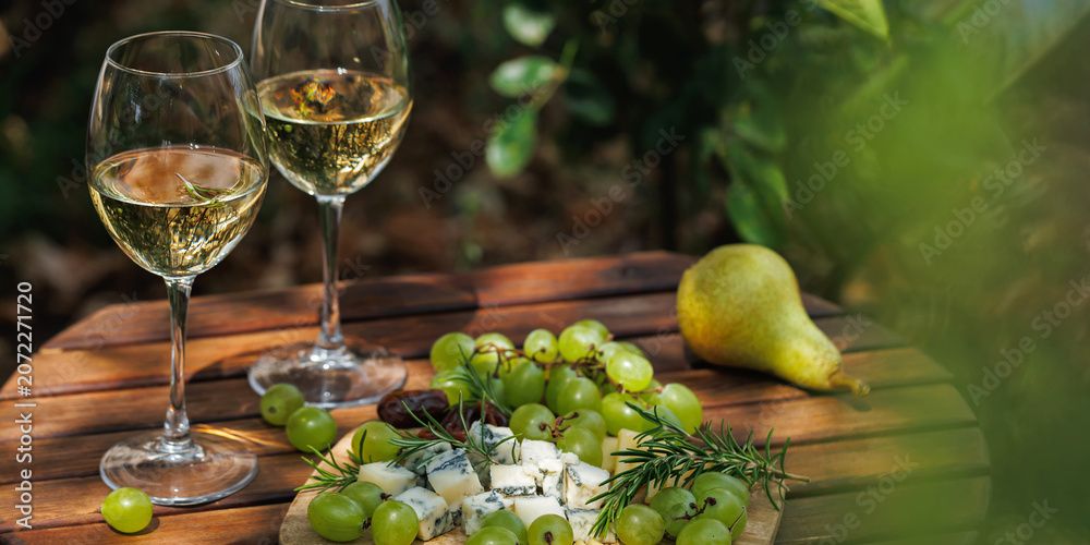 Fototapeta premium White wine glasses with cheese board grapes pear and rosemary on rustic table, sunny garden picnic, gourmet food pairing and elegant lifestyle