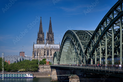 Fotografie Cologne, Germany June 22 2026: Classic view of Cologne Cathedral and Hohenzollern Bridge with love locks and pedestrians on a sunny day in Germany
