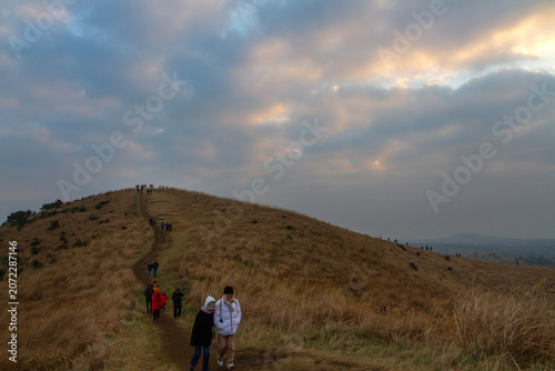 Konstfotografi Hikers ascend a grassy hill under a dramatic cloudy sky at sunset