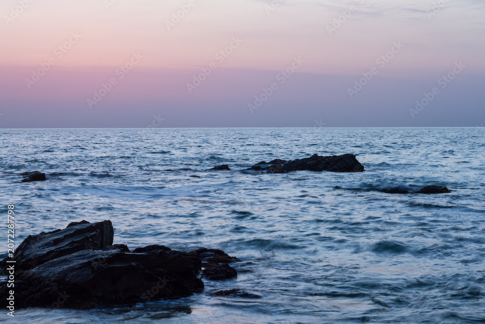 Obraz premium Serene ocean waves gently lap against dark rocks at twilight with a soft pink and purple sky