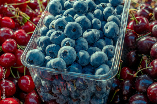 Fresh organic blueberries in plastic container surrounded by sweet red cherries at local farmers market grocery store summer berries background