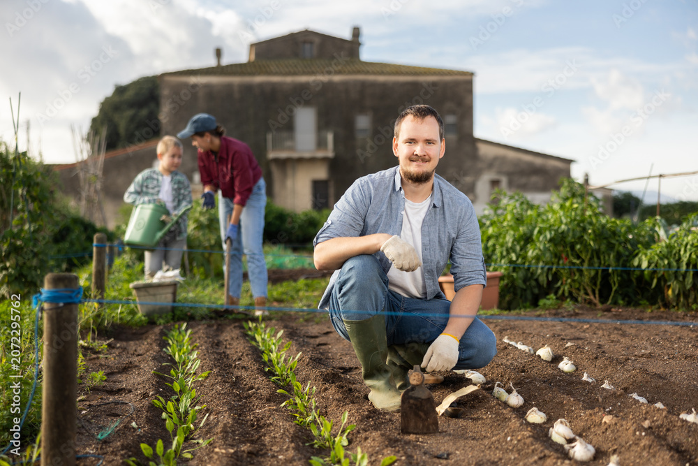 Obraz premium Male farmer plants garlic in the garden with his family on the background. Farmer has planted and grows organic seeds.