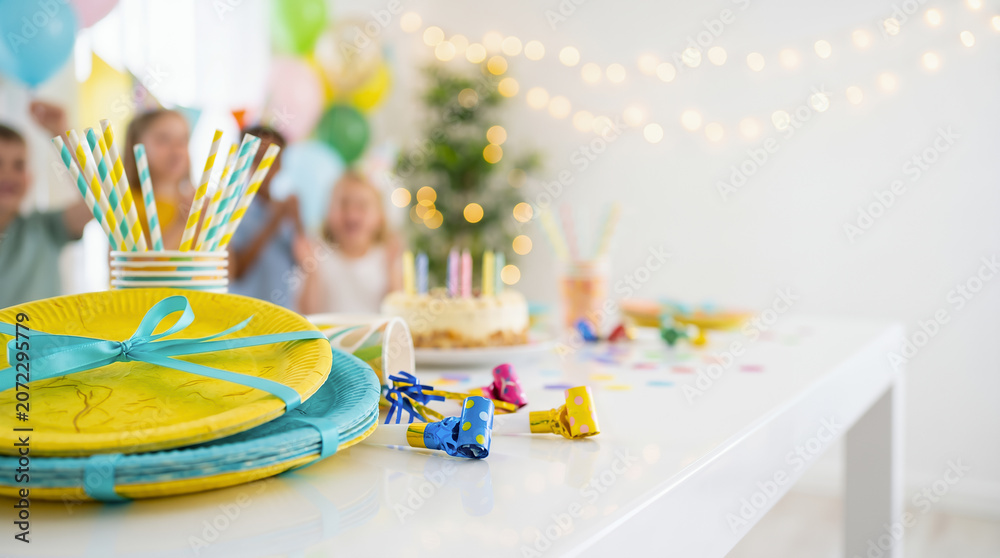 Fototapeta premium Party table with yellow plates, striped straws and polka dot blowers. Blurred children celebrating and cake with candles background. Happy birthday card concepts. Celebration banner with copy-space.