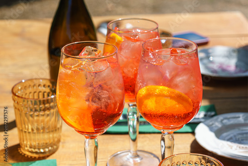 Three glasses of refreshing orange spritz cocktails with ice and citrus slices served outdoors on green sunny patio. Summer relaxation, outdoor dining, leisure, travel and social gathering concept.
