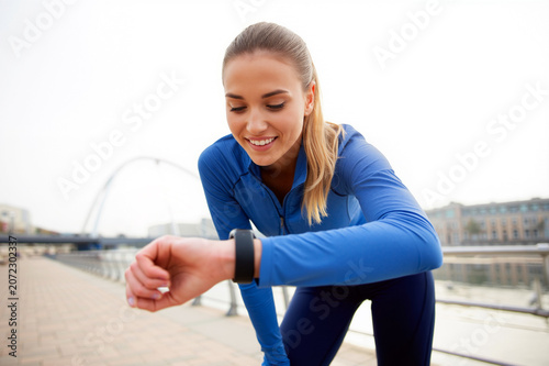 A happy young woman in blue sportswear monitors her heart rate and workout progress on a smartwatch during a morning jog in the city. Generative AI.