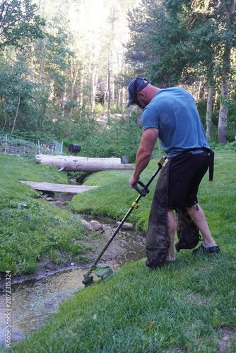 Authentic documentary-style image of a man trimming vegetation along a mountain creek while wearing protective workwear. Real-life outdoor maintenance highlighting environmental stewardship, sustainab