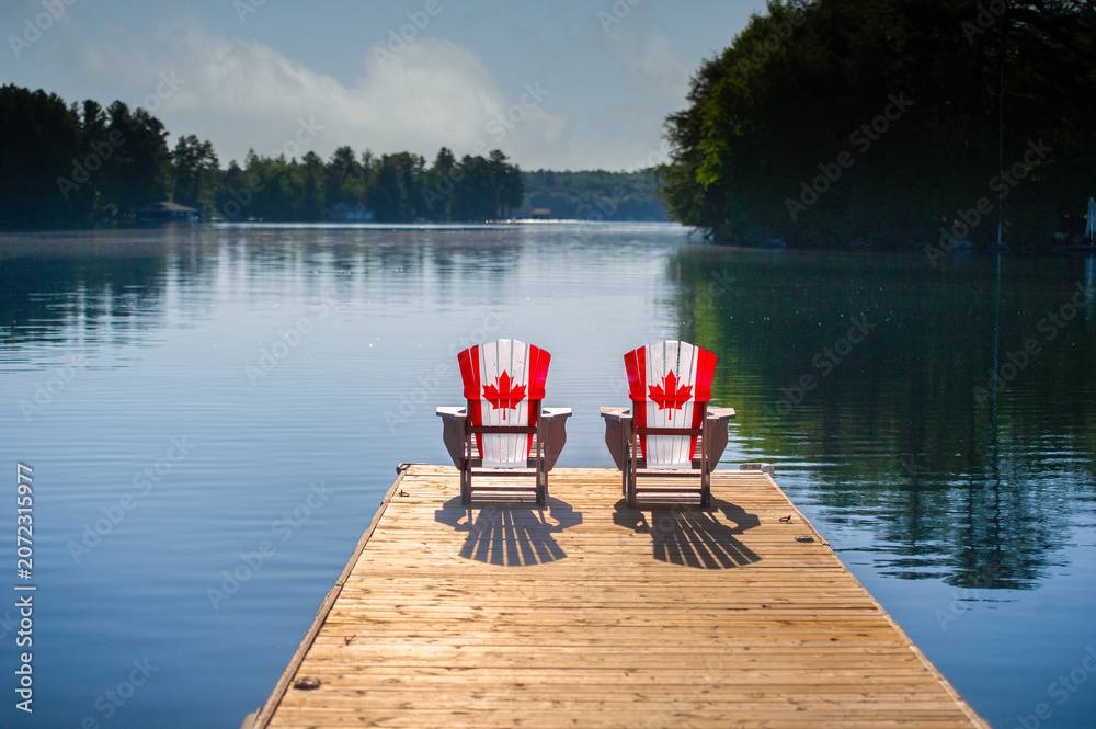 Fototapeta premium Two Adirondack chairs painted with the Canadian flag sit on a wooden dock overlooking a calm Muskoka lake, framed by forested shores under a soft blue summer sky. Canada Day cottage country.