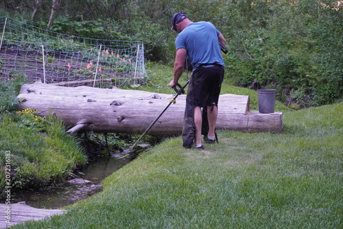Authentic documentary-style image of a man trimming vegetation along a small creek beside a fenced garden on a rural mountain property. Real-life outdoor maintenance highlighting sustainable land care