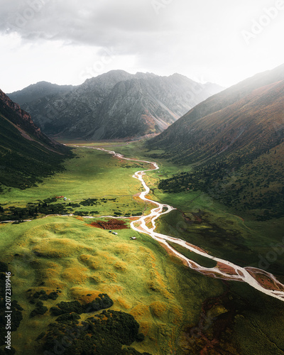 Aerial view of the winding river flowing through the lush green Turgen Valley surrounded by majestic mountains under a soft cloudy sky in Turgen, Kyrgyzstan.