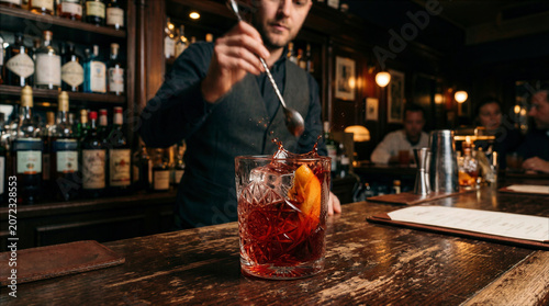 Professional bartender stirring a red cocktail with splashing liquid, ice, and orange peel garnish in a moody upscale speakeasy bar.