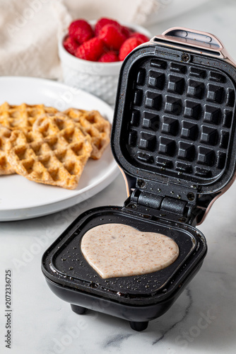 A mini waffle maker with batter cooking in the griddle in the shape of a heart.