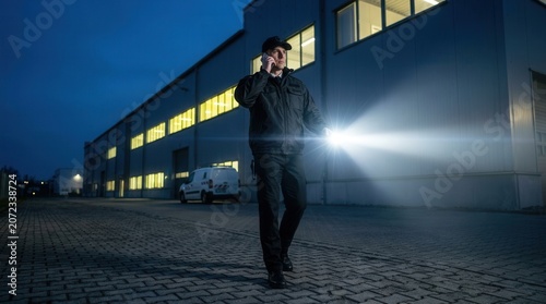 A professional security guard on patrol using a flashlight and mobile phone.