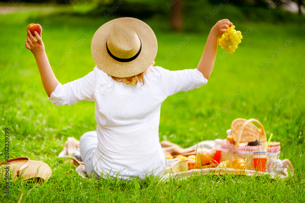 Obraz premium Happy blonde woman in straw hat with peach and grapes on picnic blanket. Raised arms, basket, summer vacation and country freedom concept, rear view from back.
