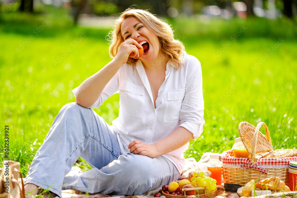 Fototapeta premium Laughing blonde woman with fresh peach on picnic blanket in park. Cheerful emotions, summer fruit snack and healthy eating concept.