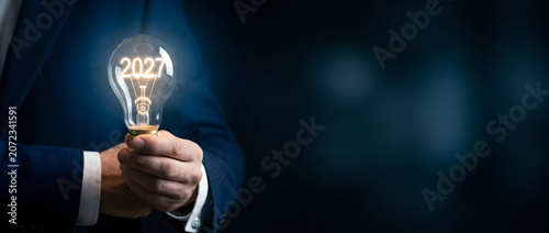 Businessman hand holding glowing light bulb with 2027 filament text on dark blue background
