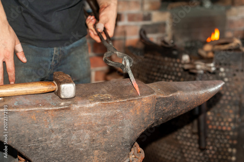 Bild auf Leinwand A forger wearing blue jeans and a T-shirt holds fire tongs and a twisted metal bolt on a vintage anvil