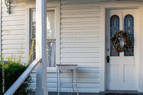 The entrance to a historic white building with a double glass window-style door. A colorful, festive Christmas wreath hangs on the vintage door. The entry has a small white table on the porch steps.