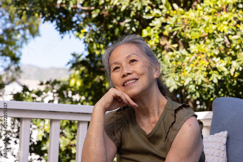Fototapeta premium Senior Asian woman sitting on porch, resting chin on hand, framed by white railing and pillow
