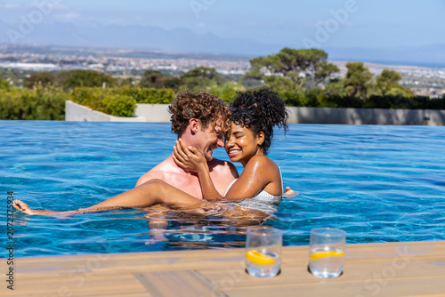 Diverse couple embracing in infinity pool at rooftop wooden deck in swimwear with two lemon glasses
