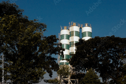 Vászonkép Industrial concrete silo plant framed by lush green trees, cement manufacturing