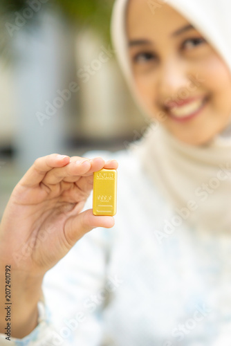 A young Asian Muslim woman is holding a gold bar. She looks very happy and joyful.