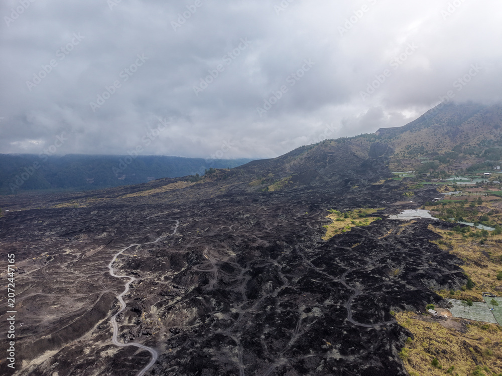 Obraz premium Aerial view of black volcanic lava fields and winding dirt tracks at the foot of Mount Batur under a dramatic cloudy sky in Bali, Indonesia