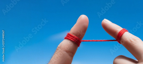 Two fingers in close-up against a sky background, tightly tied with a red thread. Symbol of an unbreakable bond, love, and destiny. Concept of relationships, marriage, and spiritual union of two peopl