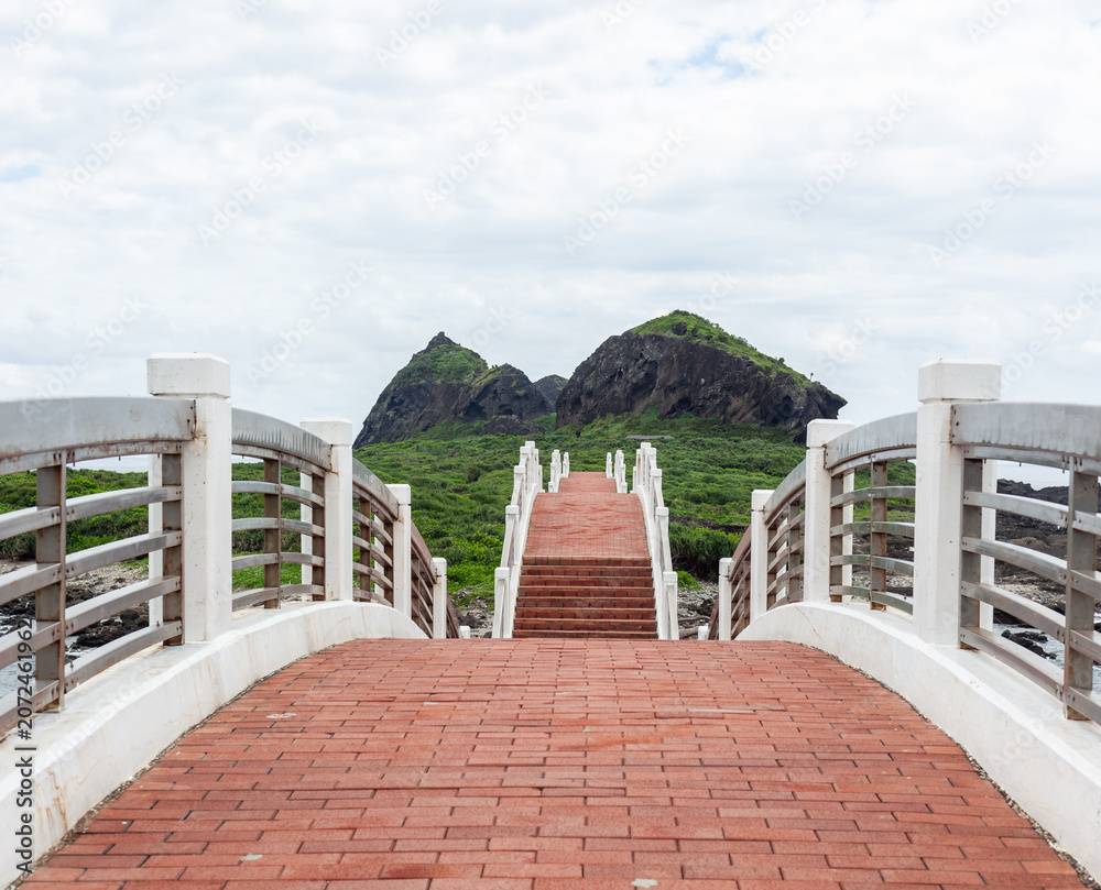 Obraz premium Sanxiantai Arch Bridge in Taiwan. Red brick pathway leads to volcanic rock formations. Lush green hills under a cloudy sky. Coastal walkway view.