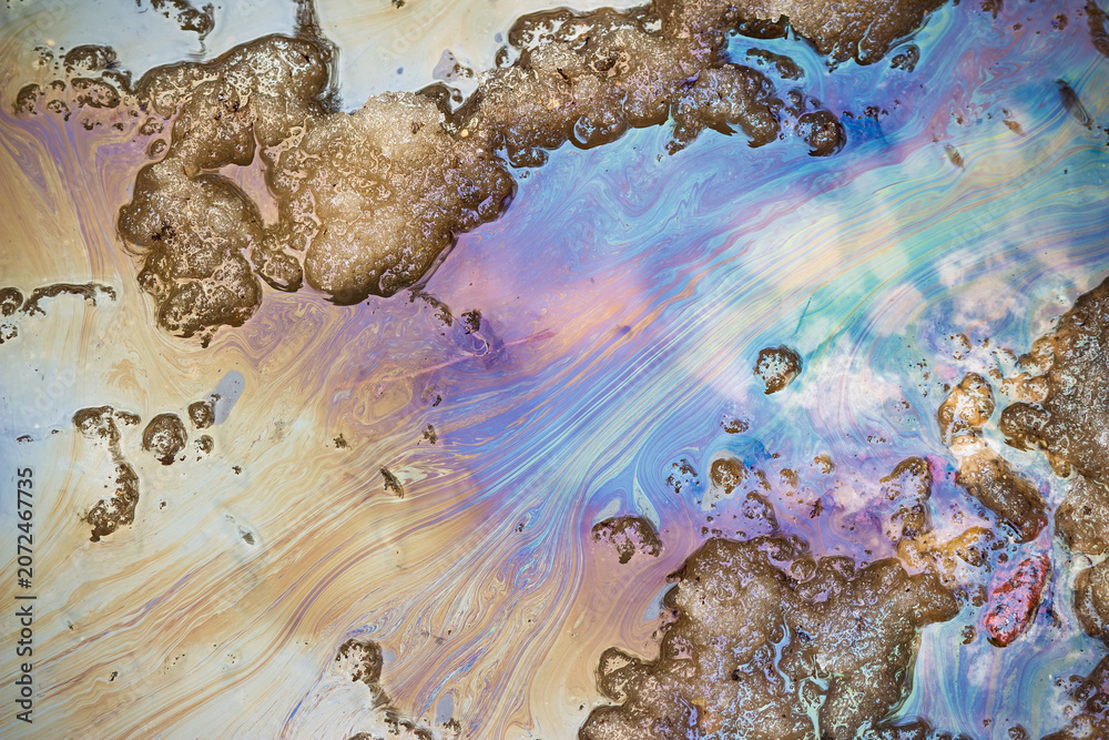 Naklejka premium Iridescent oil slick spreading on a muddy spring puddle with melting snow, showing vivid gasoline stains and toxic chemical pollution pattern on the asphalt road surface