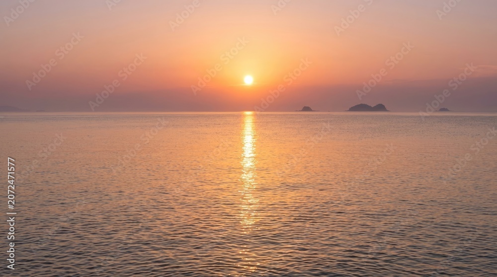 Fototapeta premium The sun sets over a calm ocean, casting a brilliant golden ray across the water. Silhouetted islands appear in the distance, adding depth to the serene scene.