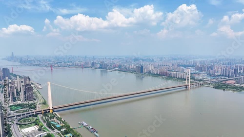 Aerial timelapse of Yangsigang Yangtze River Bridge in Wuhan, Hubei China, sunny day with blue sky and white clouds, city skyline along Yangtze river, golden double-deck suspension bridge, urban river