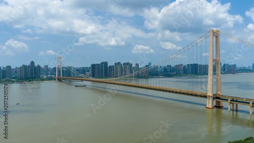 Aerial timelapse of Yangsigang Yangtze River Bridge in Wuhan, Hubei China, sunny day with blue sky and white clouds, city skyline along Yangtze river, golden double-deck suspension bridge, urban river