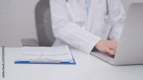 Professional doctor wearing lab coat and stethoscope using laptop computer and taking report notes on notebook at desk in hospital clinic office, medical technology and healthcare concept