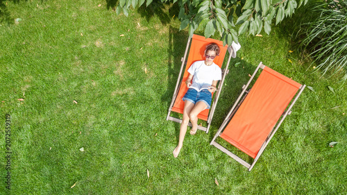 Young girl relaxes and reading book in summer garden in sunbed deckchair on grass, relaxing outdoors in green park on weekend, aerial drone view from above