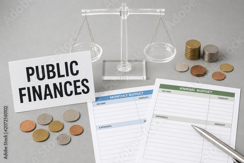 Public finances concept with balance scale, coins, monthly and annual budget sheets, and pen on a gray desktop.