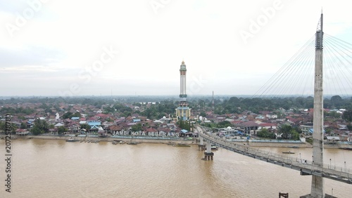 Aerial view of Jembatan Gentala Arasy spanning the Batang Hari River with the Jambi Kota Seberang district and historic rooftops in Kecamatan Jambi Timur, Jambi, Indonesia.