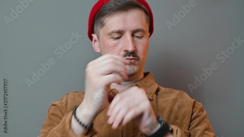 A man of middle age adjusts his wrist while wearing a red beanie and a brown shirt. He is in a simple indoor setting with gray walls, focusing on his movements