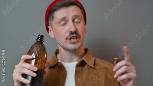 A man in his 30s with light skin drinks from a bottle while looking at the camera. He then holds up the bottle and poses, showing his playful side against a simple backdrop