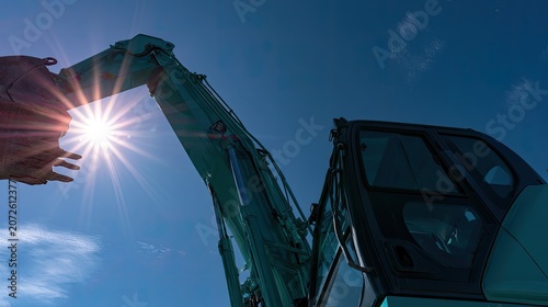Teal excavator boom and bucket angled towards a bright, starburst sun