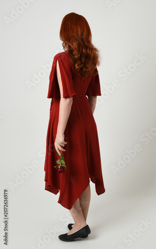 Full length portrait of beautiful redhead model wearing elegant red modern dress. standing pose, walking away facing backwards, holding a romantic red rose, isolated figure studio background