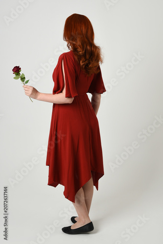 Full length portrait of beautiful redhead model wearing elegant red modern dress. standing pose, walking away facing backwards, holding a romantic red rose, isolated figure studio background