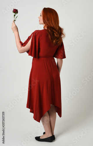 Full length portrait of beautiful redhead model wearing elegant red modern dress. standing pose, walking away facing backwards, holding a romantic red rose, isolated figure studio background