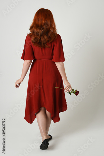 Full length portrait of beautiful redhead model wearing elegant red modern dress. standing pose, walking away facing backwards, holding a romantic red rose, isolated figure studio background