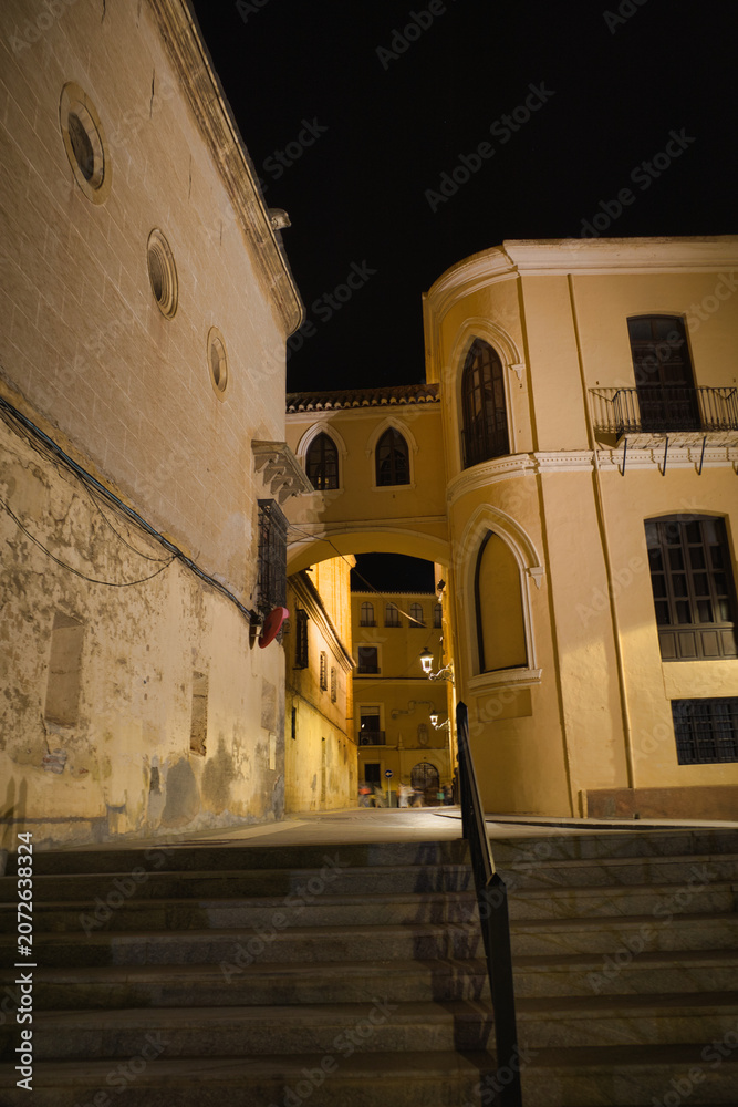 Fototapeta premium Stone steps lead upward beneath warm night lights. Ancient walls glow softly under gentle illumination. Arched passageways frame historic architecture in quietude